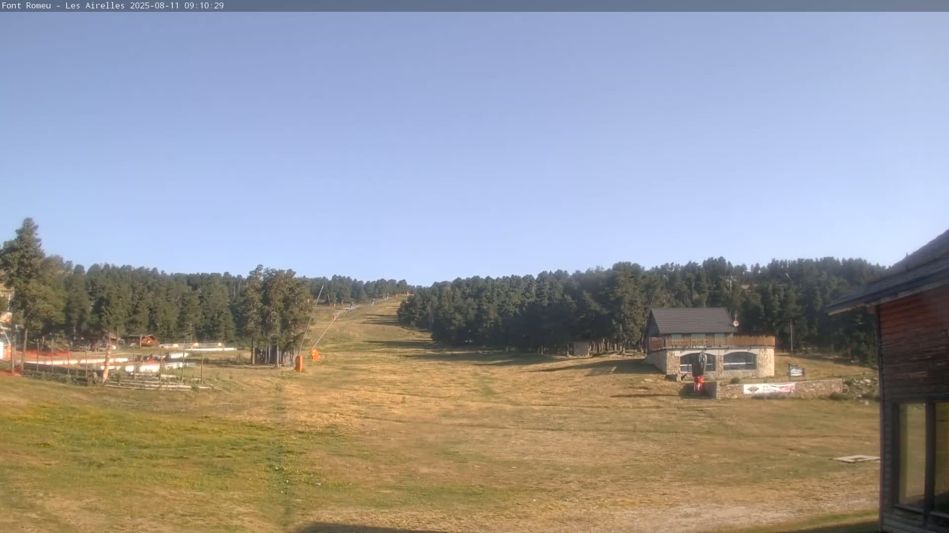 A mostly sunny day reveals a grassy ski slope flanked by evergreen trees, with a small building visible in the foreground.