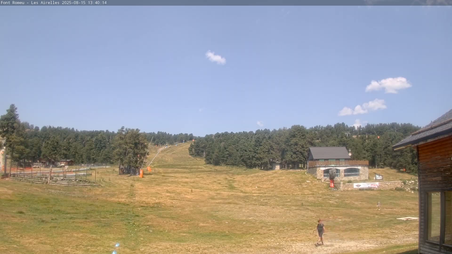 A mostly sunny day reveals a grassy ski slope flanked by evergreen trees, with a small building visible in the foreground.