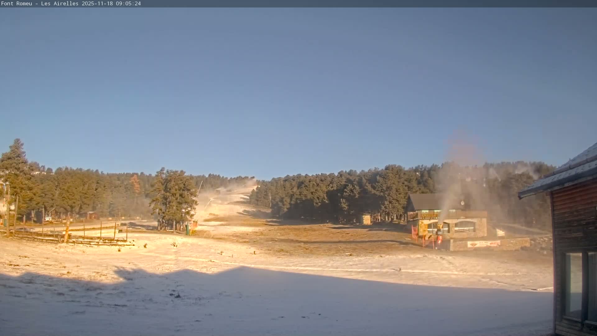 A partially snow-covered ski slope with active snow canons blowing snow onto the ground and scattered pine trees and buildings is visible under a clear, bright blue sky.