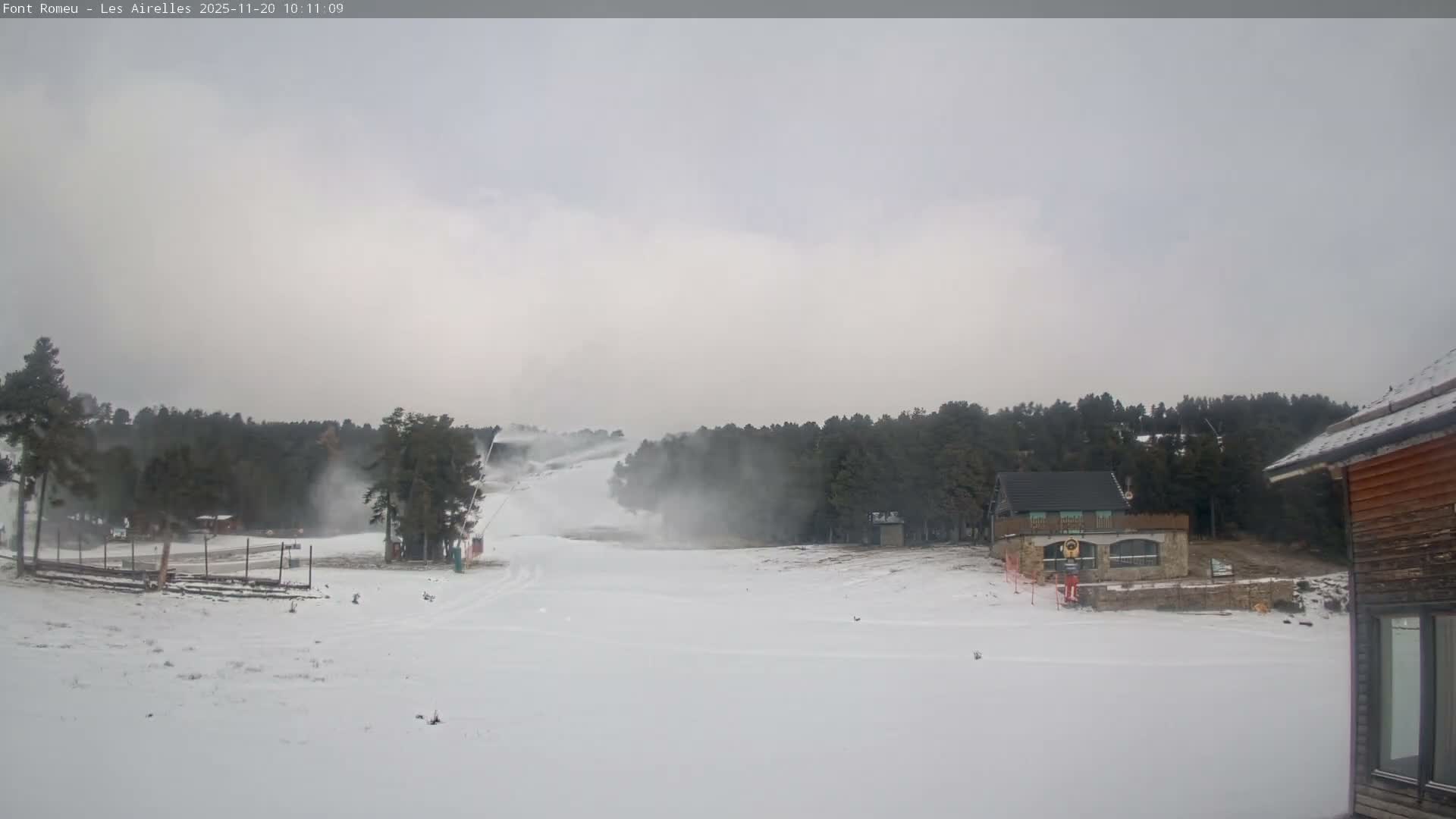 A snowy ski slope with active snow cannons creating plumes of mist and snow is visible among evergreen trees and scattered buildings, all under a heavily overcast and cloudy sky.