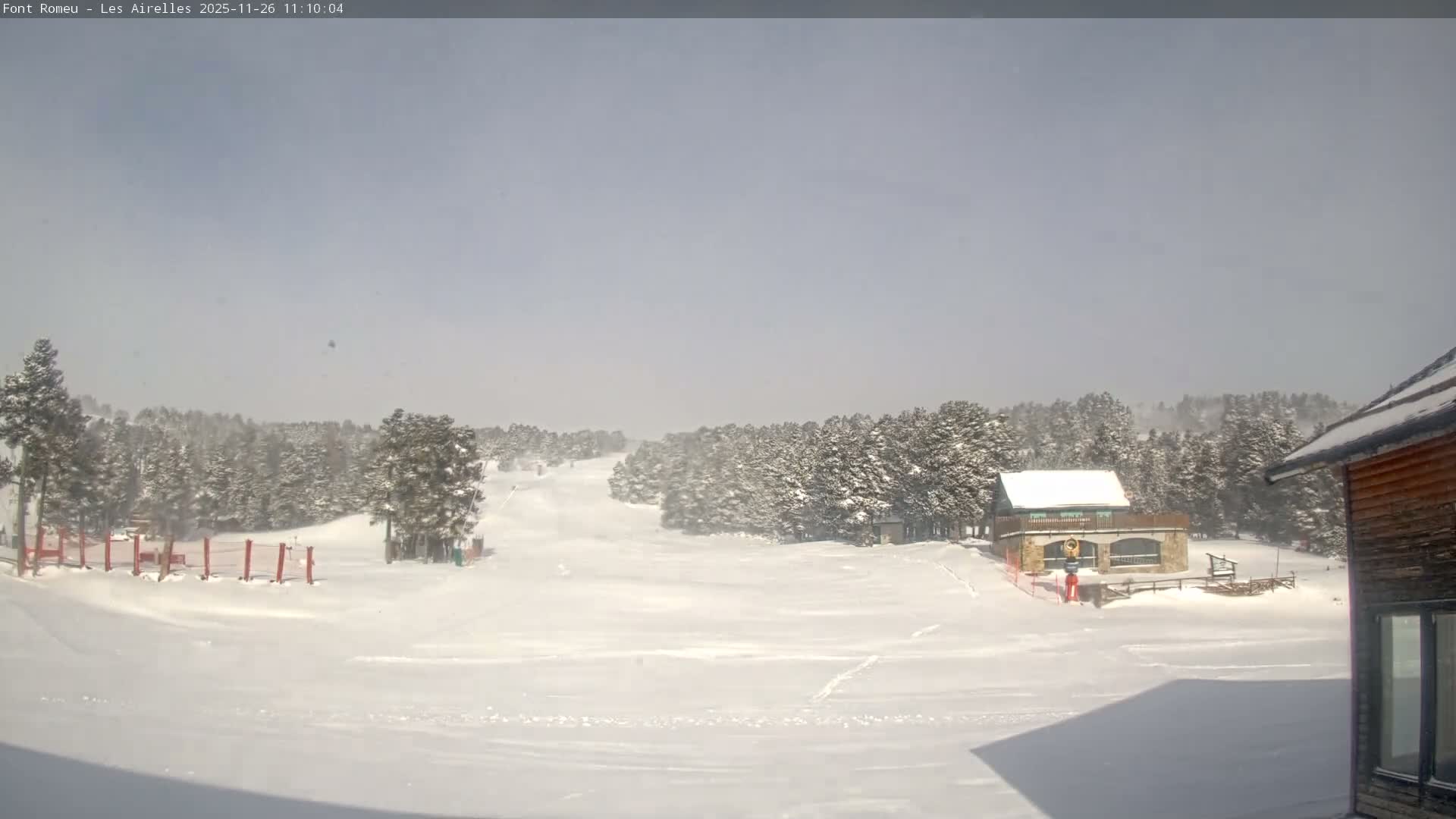 A wide, snow-covered ski slope winds through a forest of snow-laden pine trees past buildings, including a stone lodge, under an overcast sky with visible snowfall.