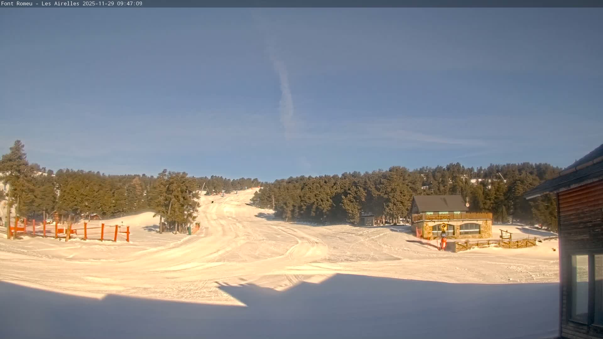 A bright winter day with clear blue skies overlooks a snowy ski slope bordered by pine forests and a stone-clad building, with long shadows cast across the foreground.