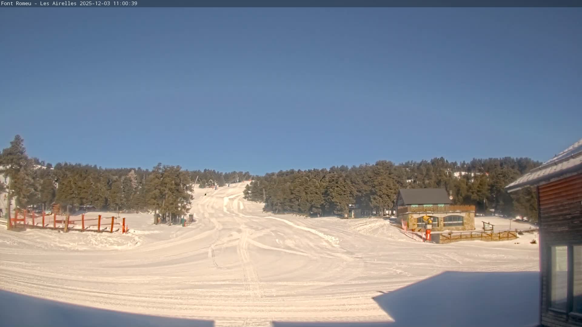 A bright, sunny day at a snowy mountain ski resort features pristine slopes with tracks, dense pine forests, and several buildings under a clear blue sky.
