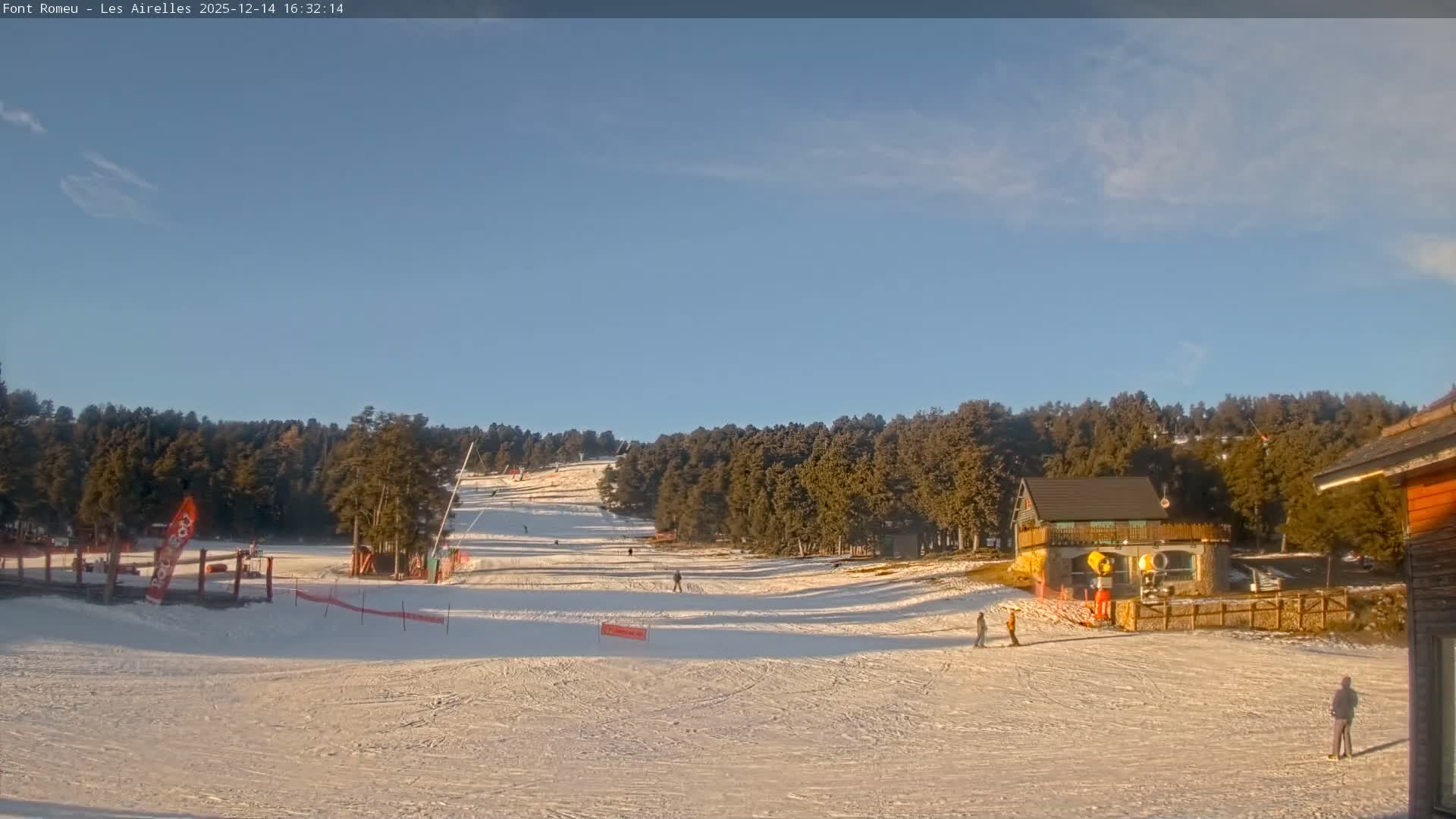 A bright, sunny day at a snowy mountain ski resort features pristine slopes with tracks, dense pine forests, and several buildings under a clear blue sky.