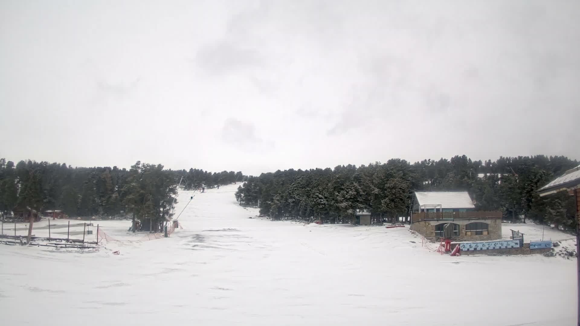 A bright, sunny day at a snowy mountain ski resort features pristine slopes with tracks, dense pine forests, and several buildings under a clear blue sky.