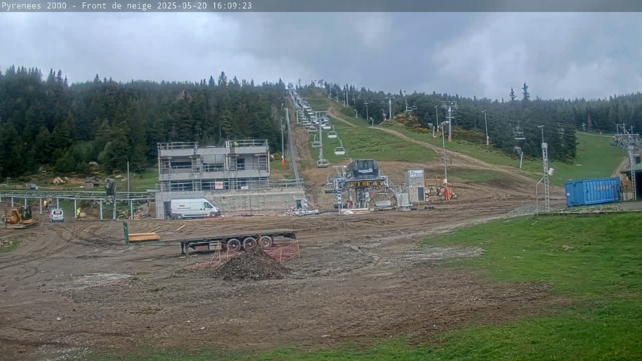 A construction site with a chairlift ascending a hill covered in green grass and evergreen trees under an overcast sky.
