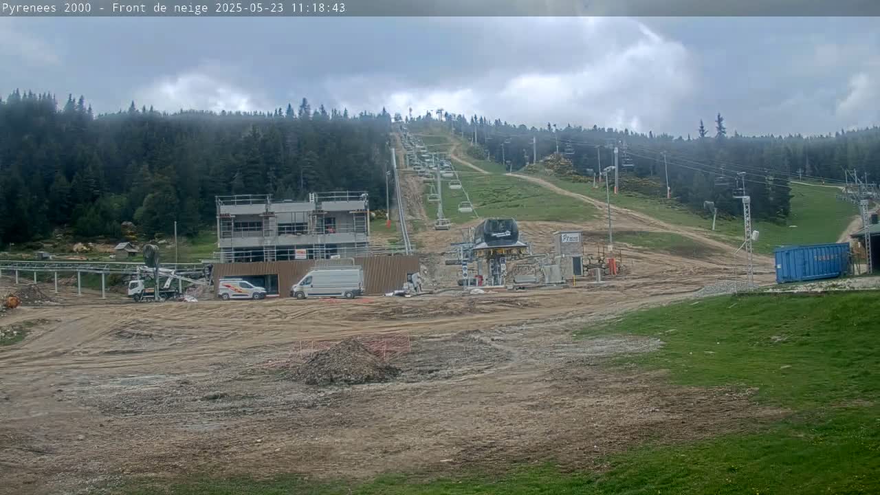 A ski lift under construction is situated on a hill surrounded by a forest under cloudy skies.