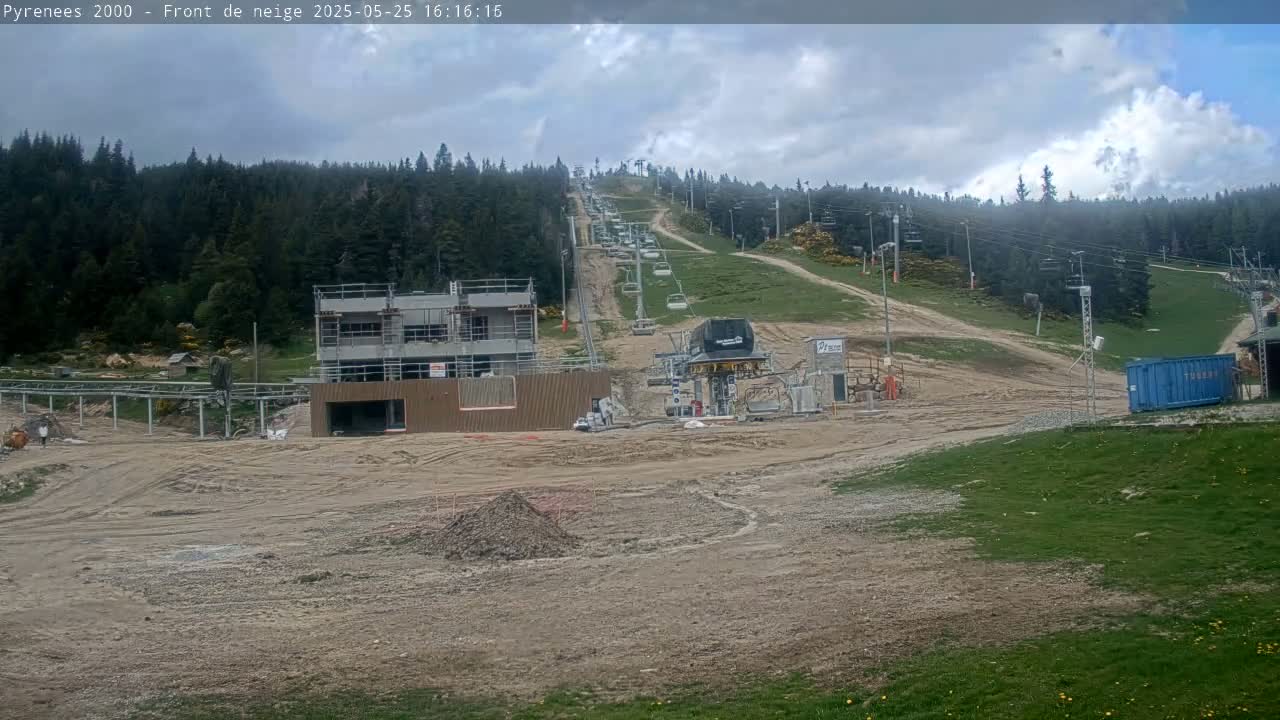 A partly cloudy day reveals a ski lift under construction in a mountainous area with a forest backdrop.