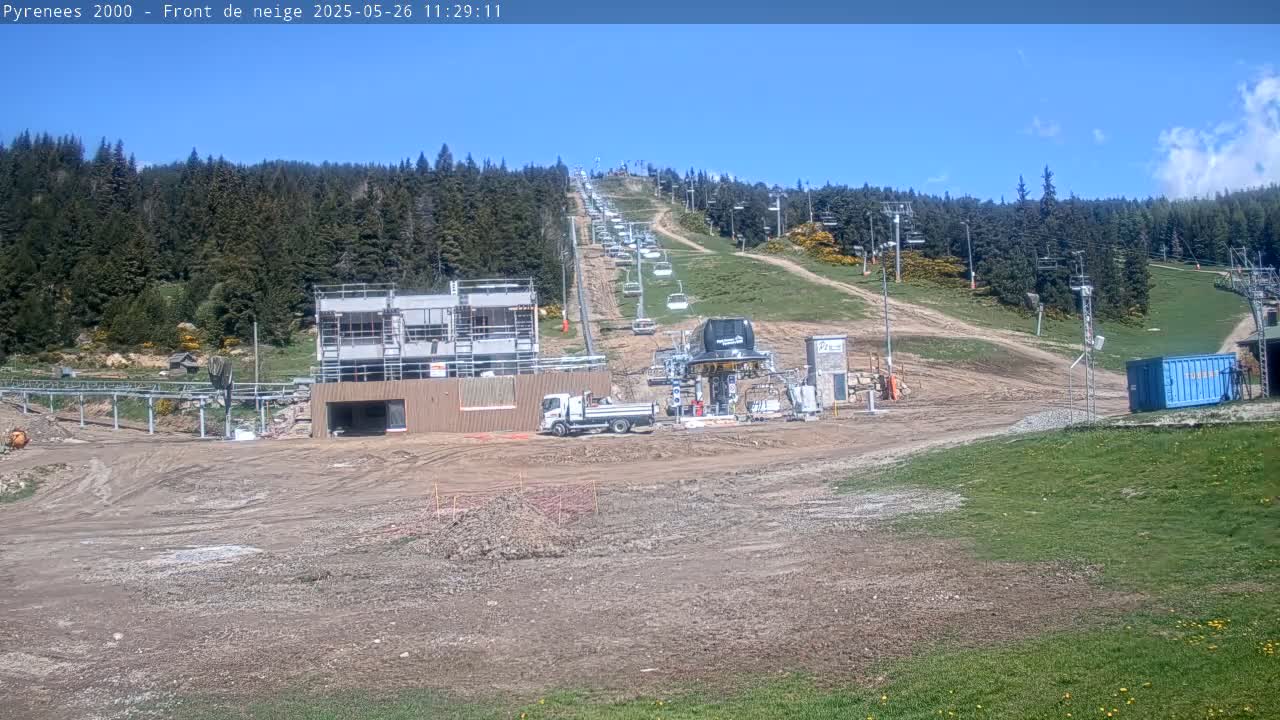 A ski lift ascends a hill covered in green vegetation and dirt, past a construction site under a clear blue sky.