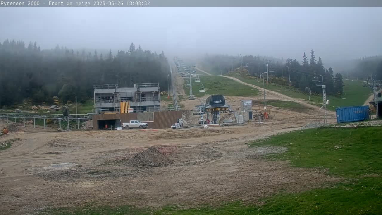 A foggy mountain ski resort shows construction in progress near a chairlift, with a mostly bare, dirt ground in the foreground and a wooded hillside in the background.