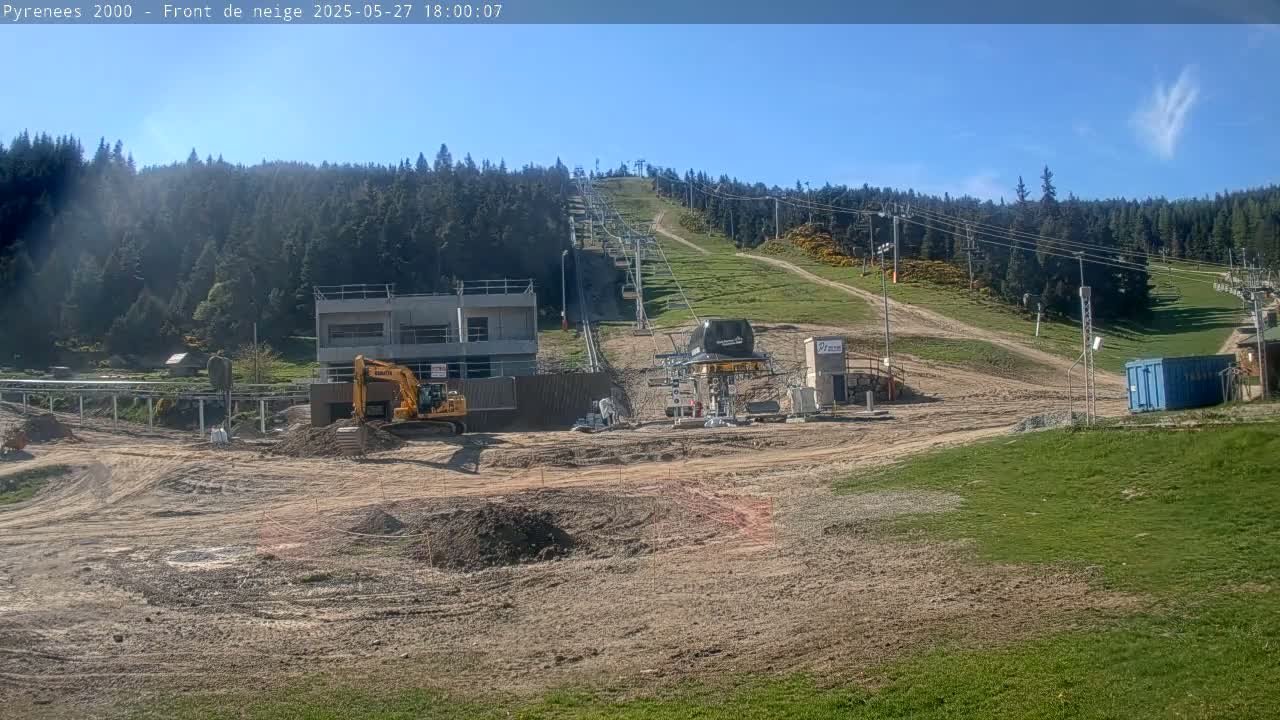 A sunny day reveals a ski lift under construction, with earthmoving equipment present on a mostly barren hillside surrounded by a coniferous forest.