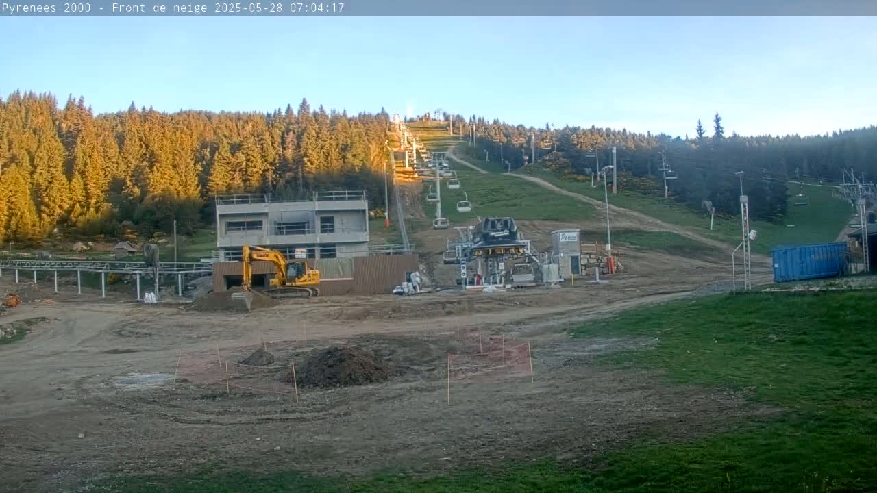 A construction site with a yellow excavator sits below a chairlift ascending a grassy hill covered in a forest of evergreens under a clear sky.