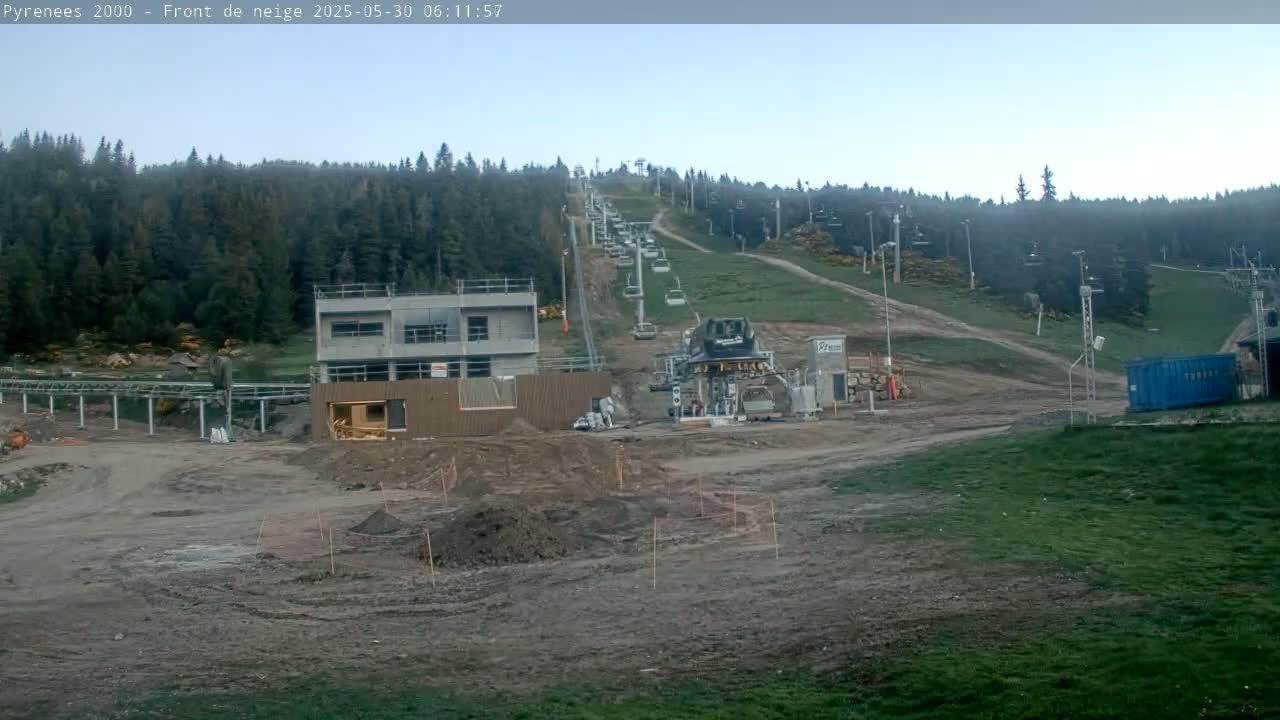 A ski lift installation is underway on a hillside covered in green grass and dark evergreen trees under a clear sky.