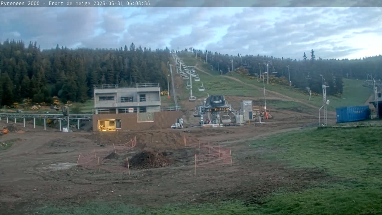 A ski lift ascends a hillside covered in green grass and coniferous trees under a cloudy sky, with construction underway at the base.
