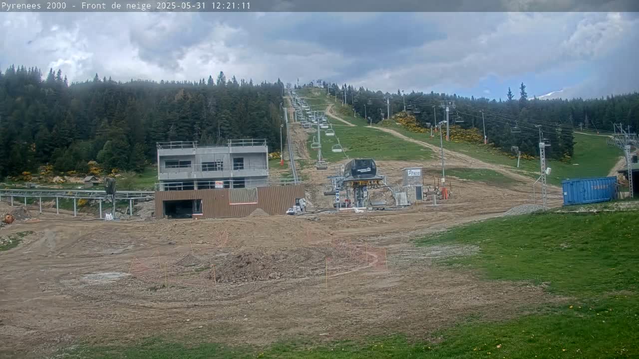 A partially cloudy sky overlooks a ski lift under construction in a mountainous area, with several chairlifts visible on the hill.