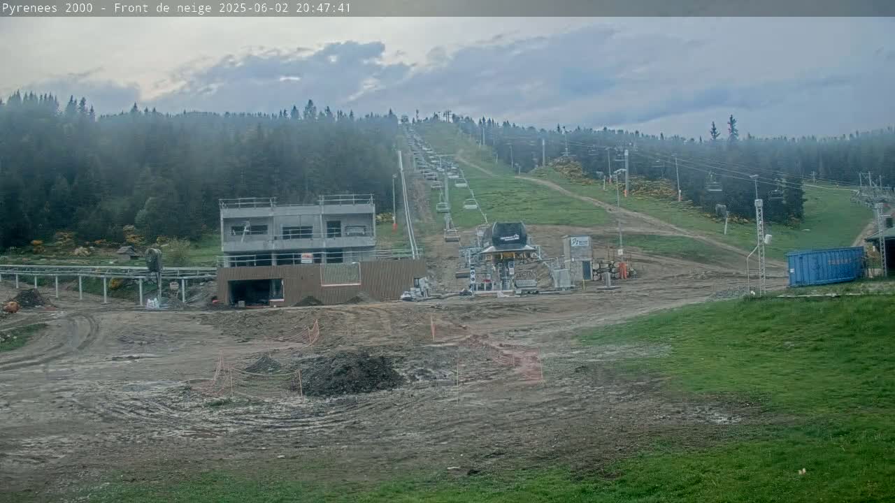 A chairlift ascends a hillside covered in a coniferous forest under an overcast sky, with muddy ground and construction visible at the bottom.
