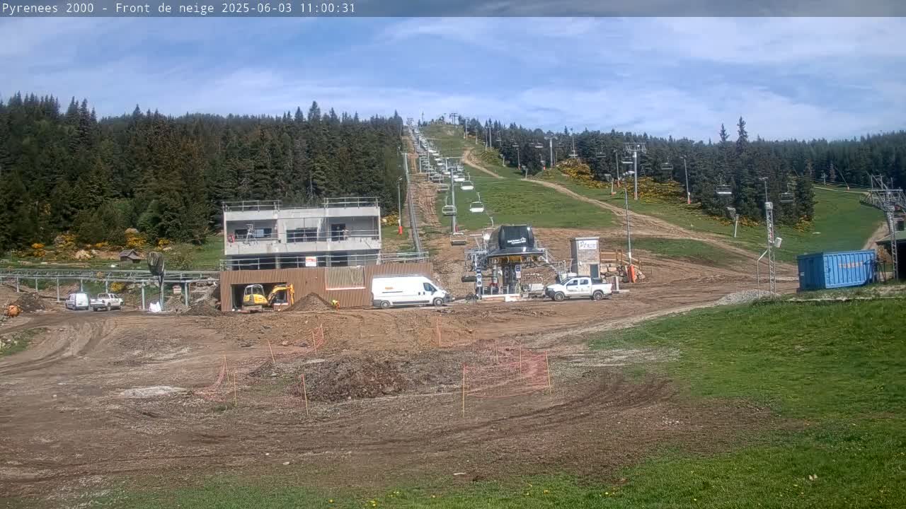 A mostly sunny day shows a ski lift ascending a hill dotted with construction equipment and vehicles amidst a mostly cleared area, surrounded by a dark green forest.