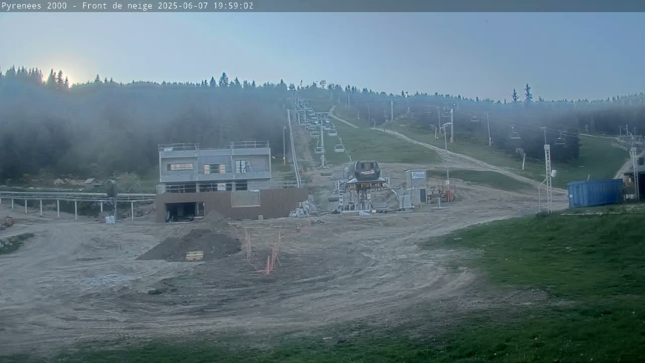 A mostly clear, hazy evening scene shows a ski lift and a partially constructed building at the base of a hill covered with a forest, with some construction work visible in the foreground.