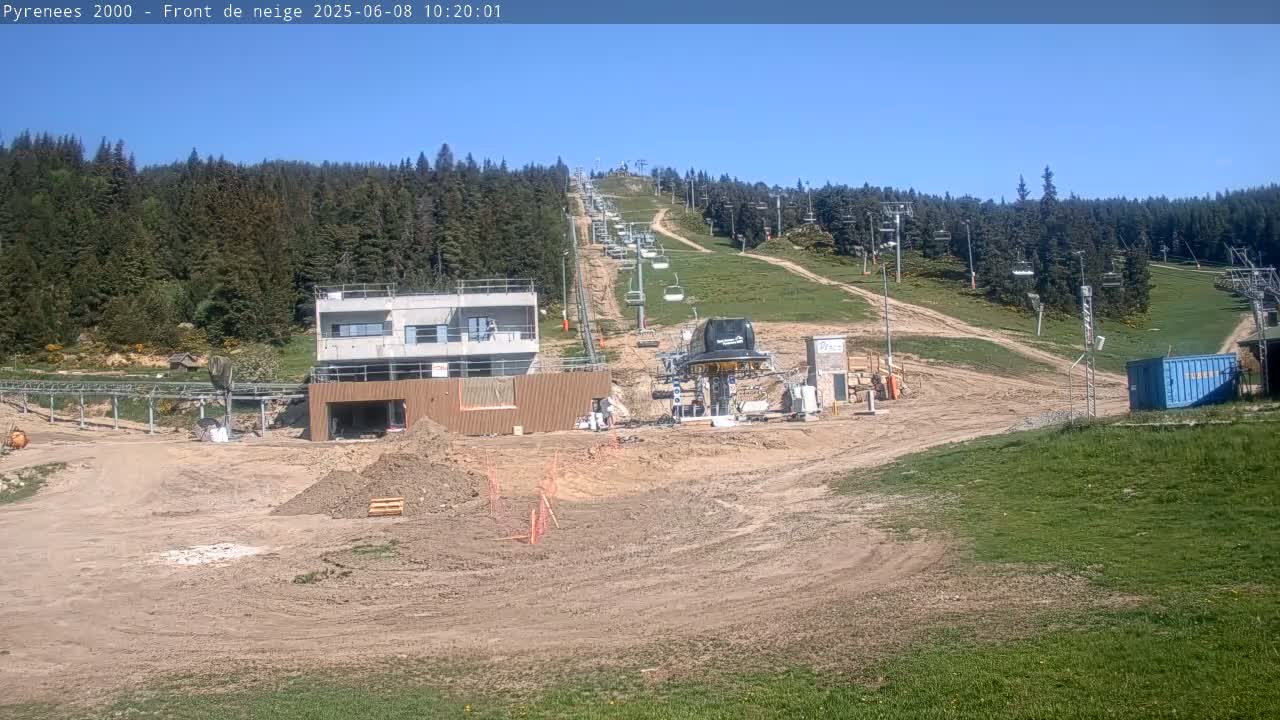 A ski lift and several buildings under construction are situated on a hillside covered in patchy grass and dirt, under a clear blue sky.