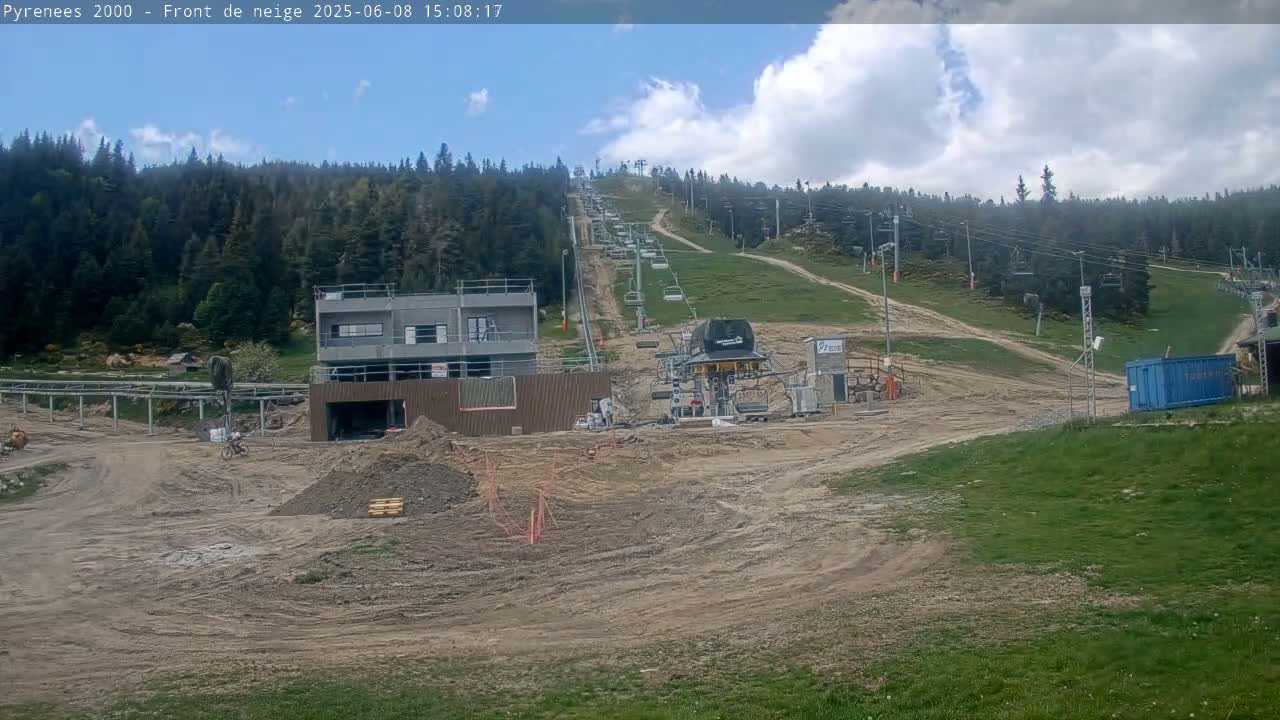 A partly cloudy day reveals a ski lift under construction on a grassy hillside surrounded by a coniferous forest.