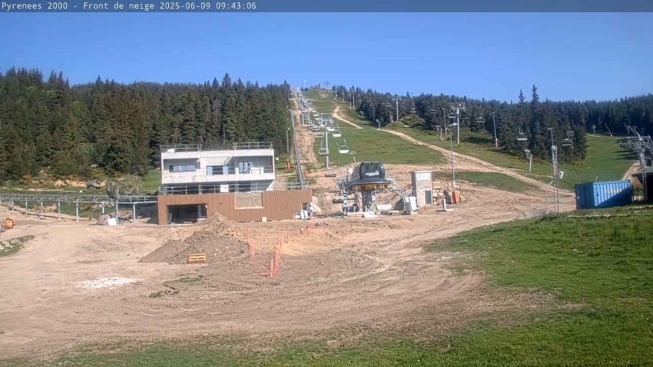 A ski lift extends uphill through a mostly clear, sunny day, past a construction site with earthworks and new buildings near the base of the mountain, surrounded by green hills and a dark forest.