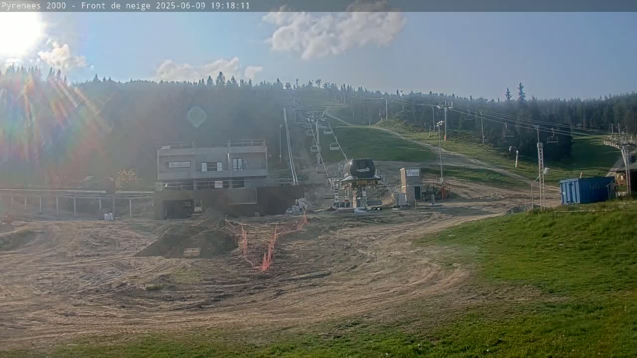 A partly cloudy, sunny day reveals a ski lift and surrounding construction area, with a background of a forested hillside.
