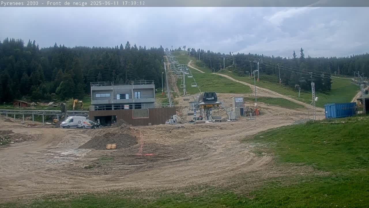 A ski lift ascends a hill under a cloudy sky, with construction taking place at the base, including a partially completed building and vehicles.