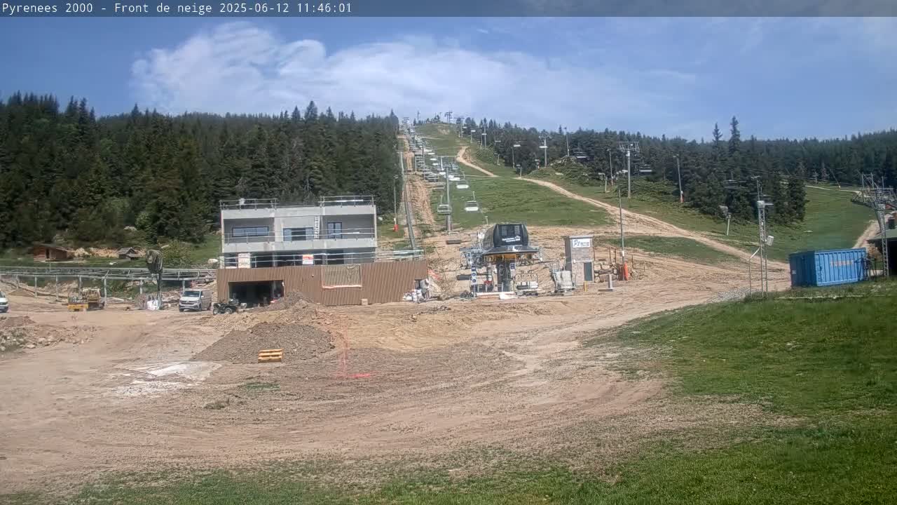 A ski lift and a partially constructed building are situated on a mostly dirt hillside, under a mostly sunny sky.
