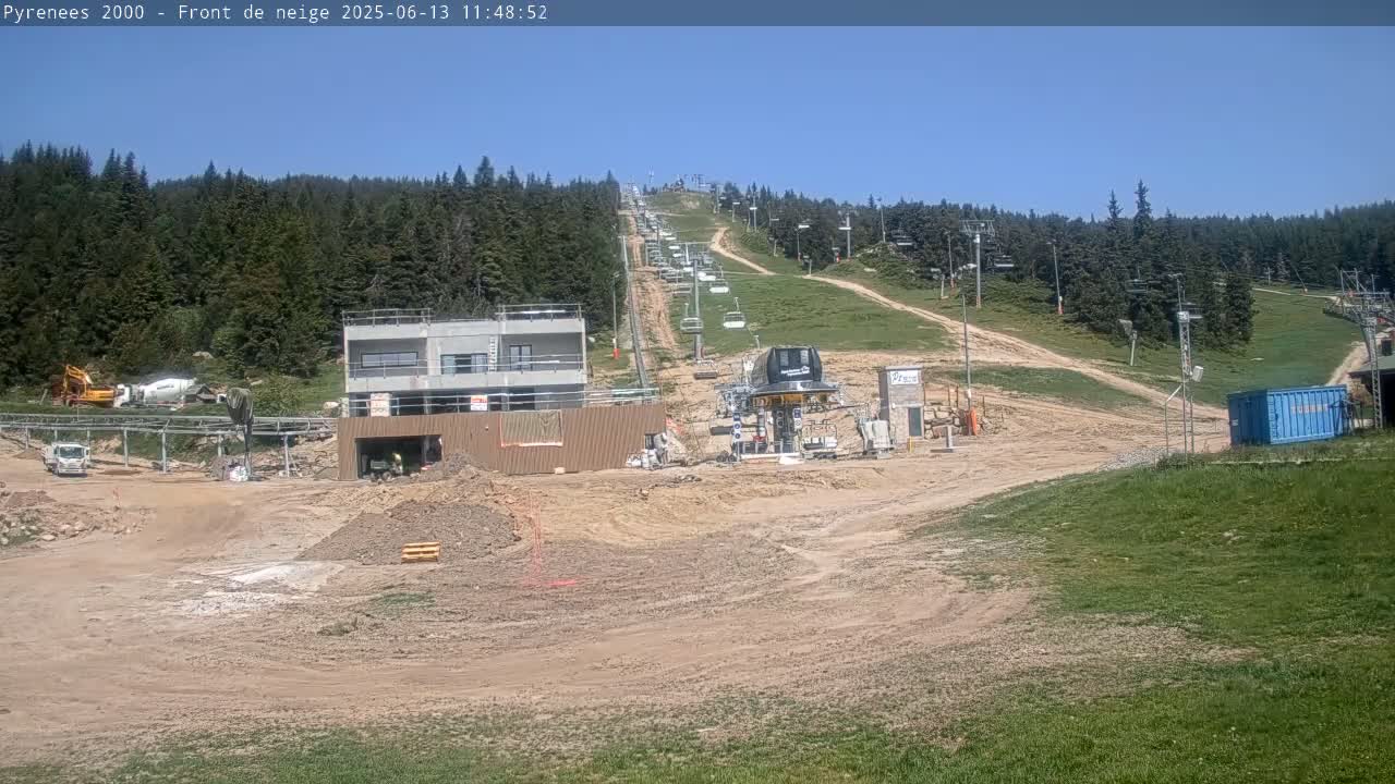 A ski lift and surrounding construction site are visible under a clear blue sky, with a forested hillside in the background.