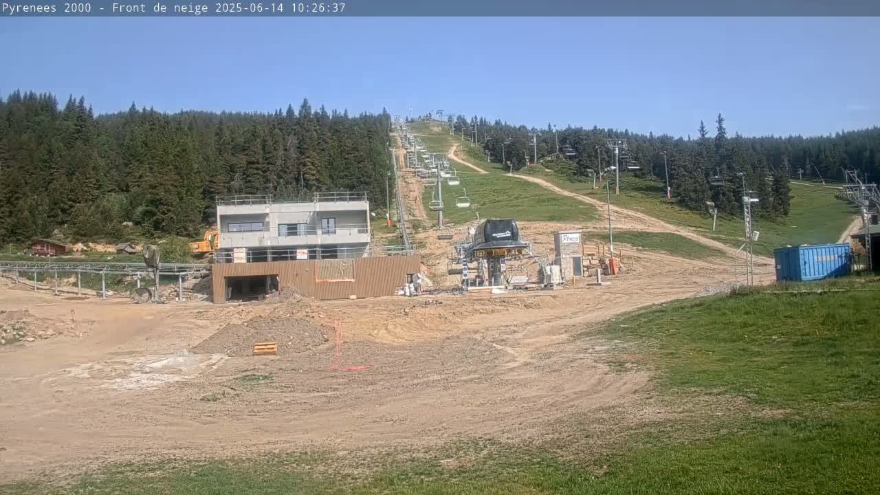A ski lift and several buildings under construction are situated on a hill surrounded by a forest under sunny skies.