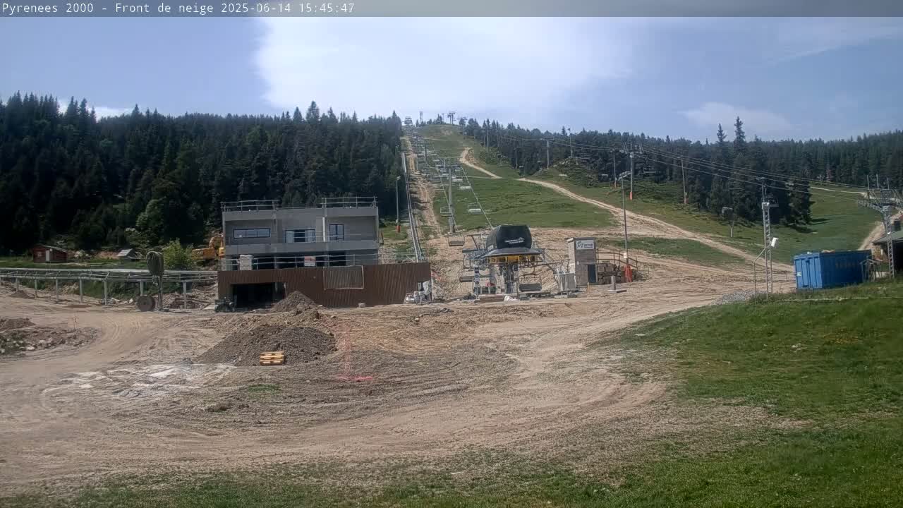 A ski lift under construction is visible on a partly sunny day, with a mostly clear sky and a forest of evergreen trees in the background.