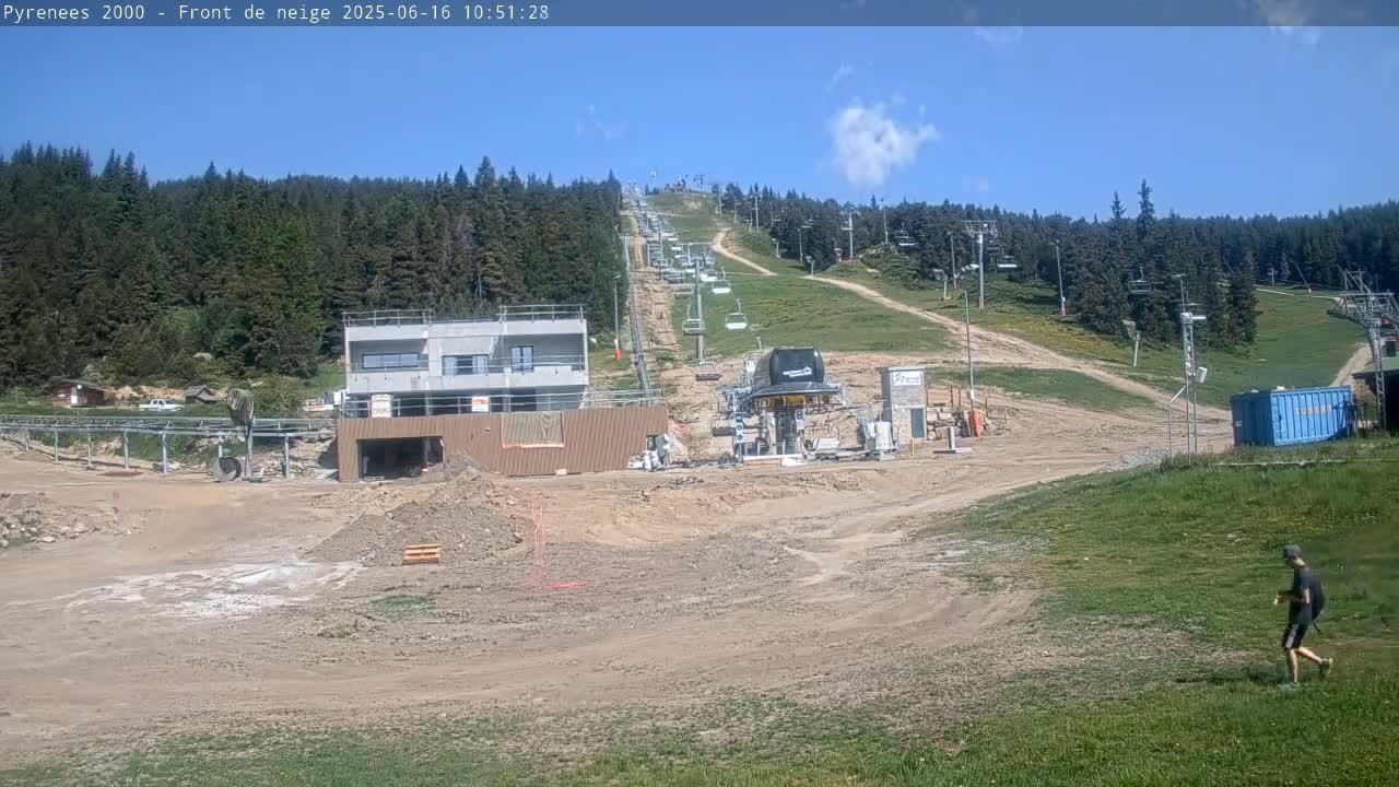 A mostly sunny day reveals a ski resort under construction, with a chairlift visible extending uphill into a green, forested mountainside.