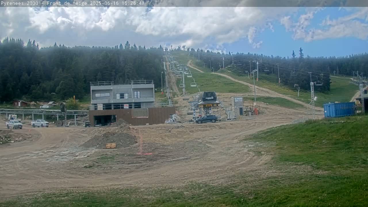 A partly cloudy day reveals a ski lift ascending a mountainside amongst construction at the base, with several vehicles parked nearby.