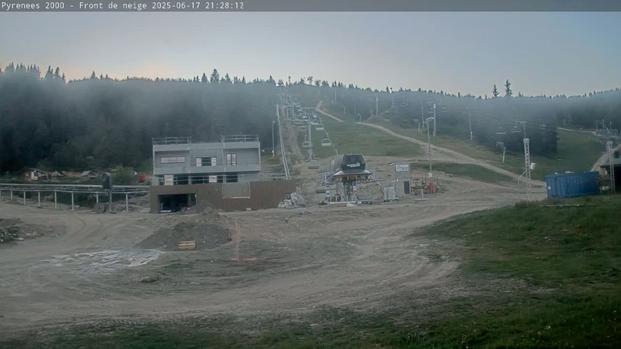 A ski lift and a building under construction are situated in a mountain valley shrouded in fog, with a dirt road leading up a grassy hillside.