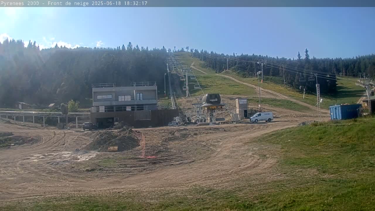 A ski lift ascends a grassy hill under a mostly clear sky, with construction underway at the base, including a building, vehicles, and earthworks.