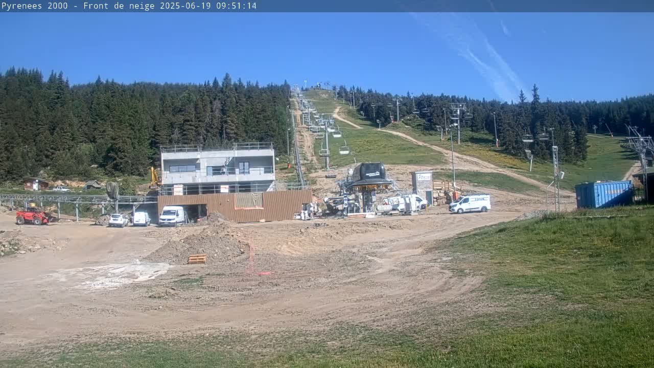A ski lift area undergoing construction is visible on a sunny day, with several vehicles parked near a partially constructed building and piles of dirt.