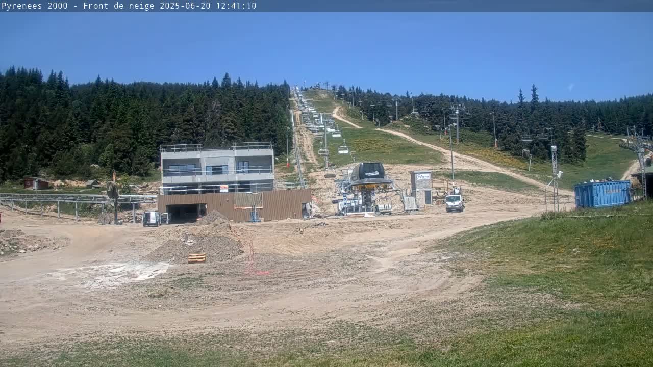 A ski lift and a partially constructed building are situated on a dirt-covered hillside, under a clear blue sky, surrounded by a coniferous forest.