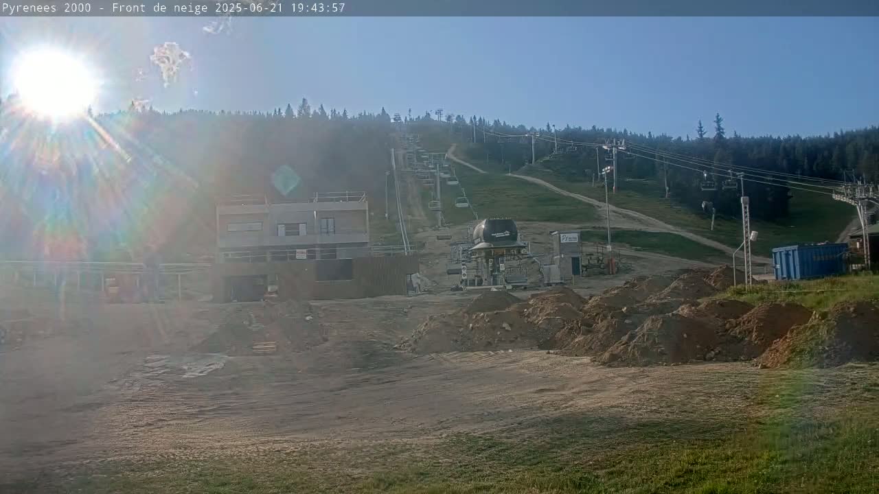 A sunny, mostly clear day reveals a ski lift ascending a grassy hillside, with construction underway at the base, featuring piles of dirt and a partially built structure.
