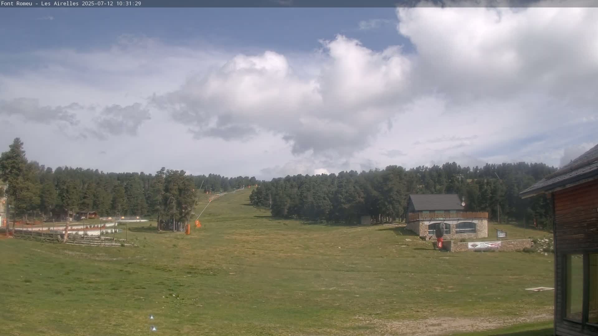A partly cloudy sky overlooks a grassy field, a ski slope, a small stone building, and a dense forest of evergreen trees.
