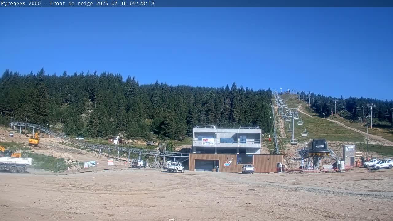 A ski lift and construction equipment are visible in a sunny, mountainous area covered in a dense evergreen forest.