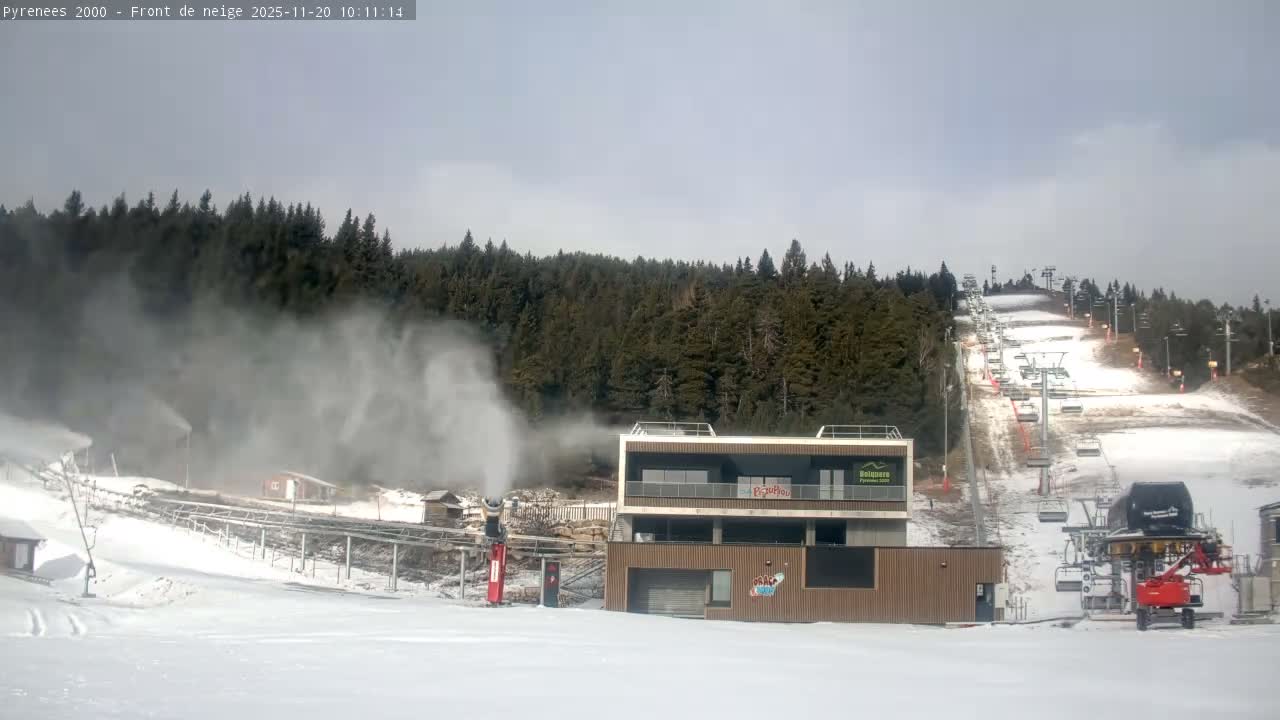 Multiple snow cannons are actively creating powder on a ski slope featuring chairlifts and a modern building, all set against a backdrop of dense evergreen forests under a dull, overcast sky.