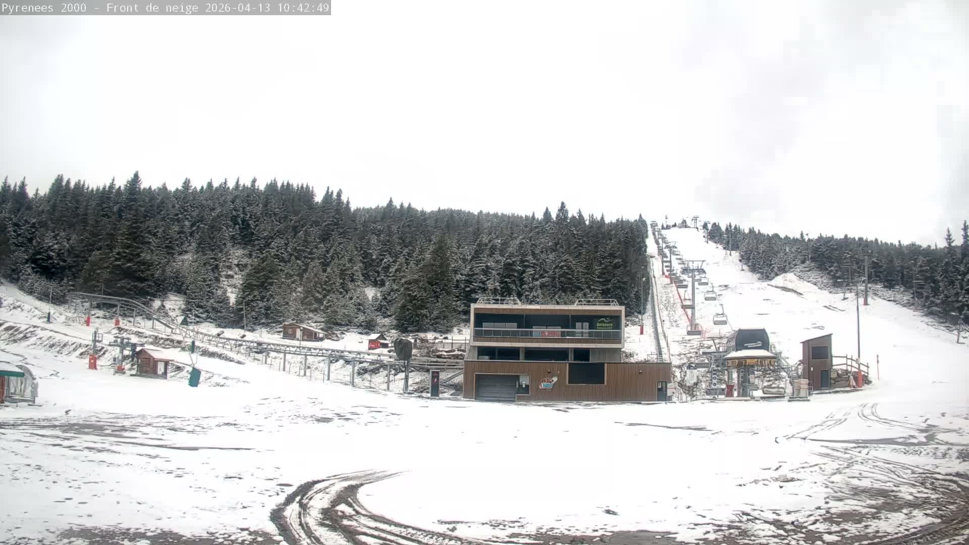 A snow-covered ski resort features a ski slope with multiple chairlifts ascending a pine-forested mountain, a modern lodge at its base, and partly cloudy skies transitioning from clear blue to overcast.