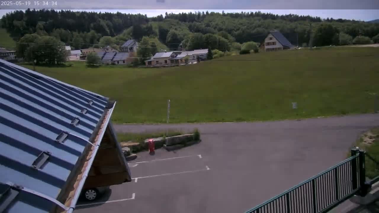 A partly cloudy day reveals a view of a parking area, a metal roof, and a small village nestled in a green valley surrounded by a forest.