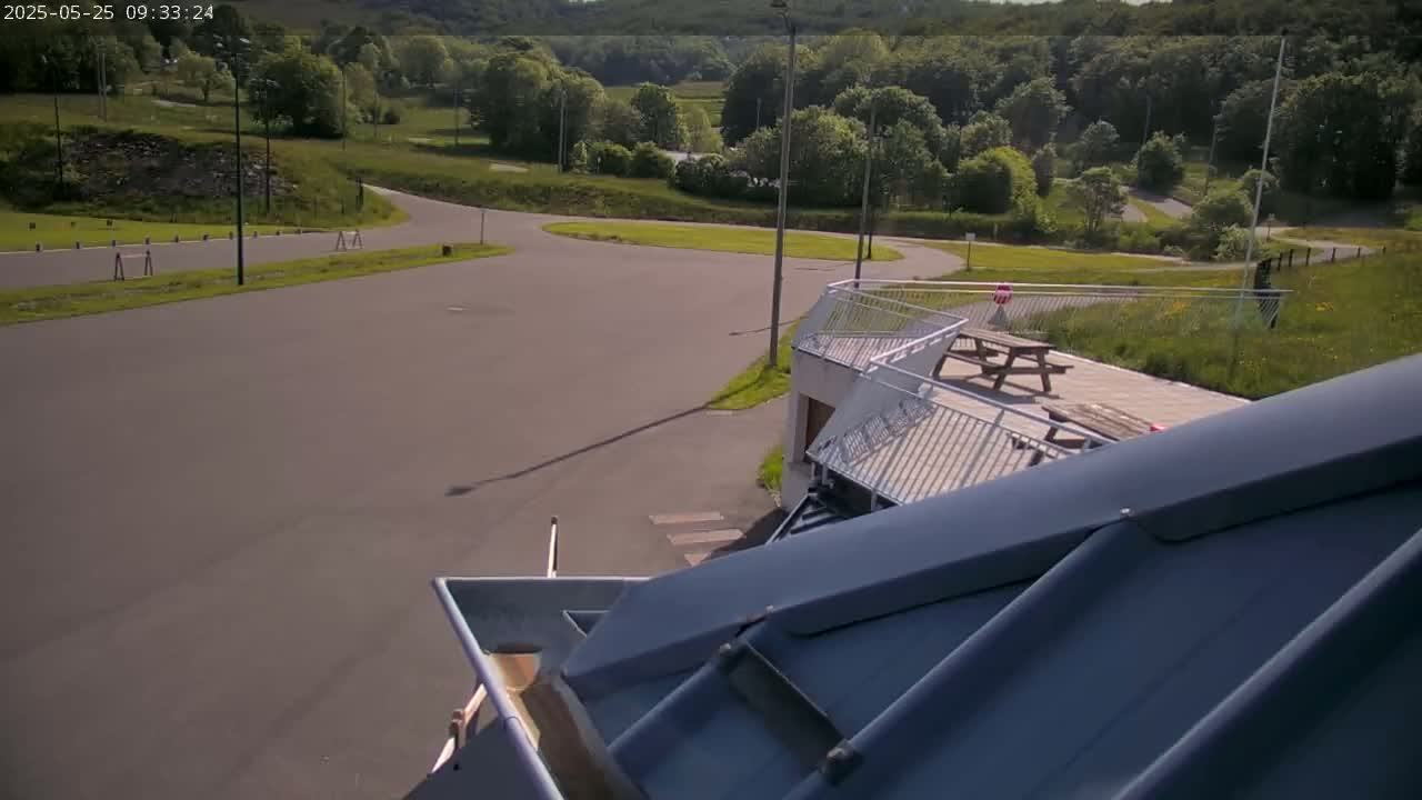 A paved area with a curving roadway, surrounded by green hills and trees under a sunny sky, is partially visible from a high vantage point above a sloped, gray roof.