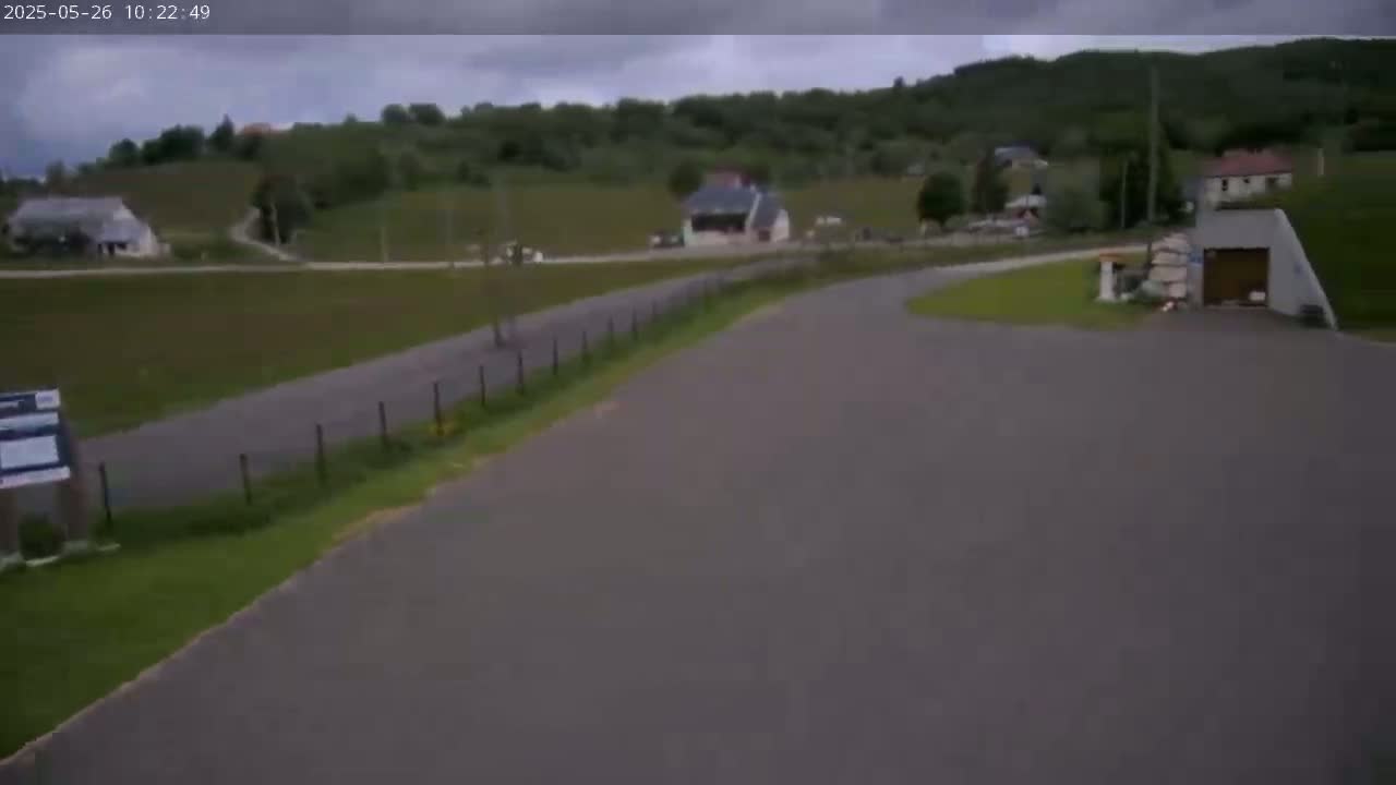 A paved road curves through a rural area with green hills and a few buildings under an overcast sky.
