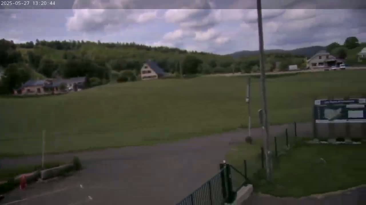 A partly cloudy day reveals a grassy field with several houses nestled among green hills and trees in the background.