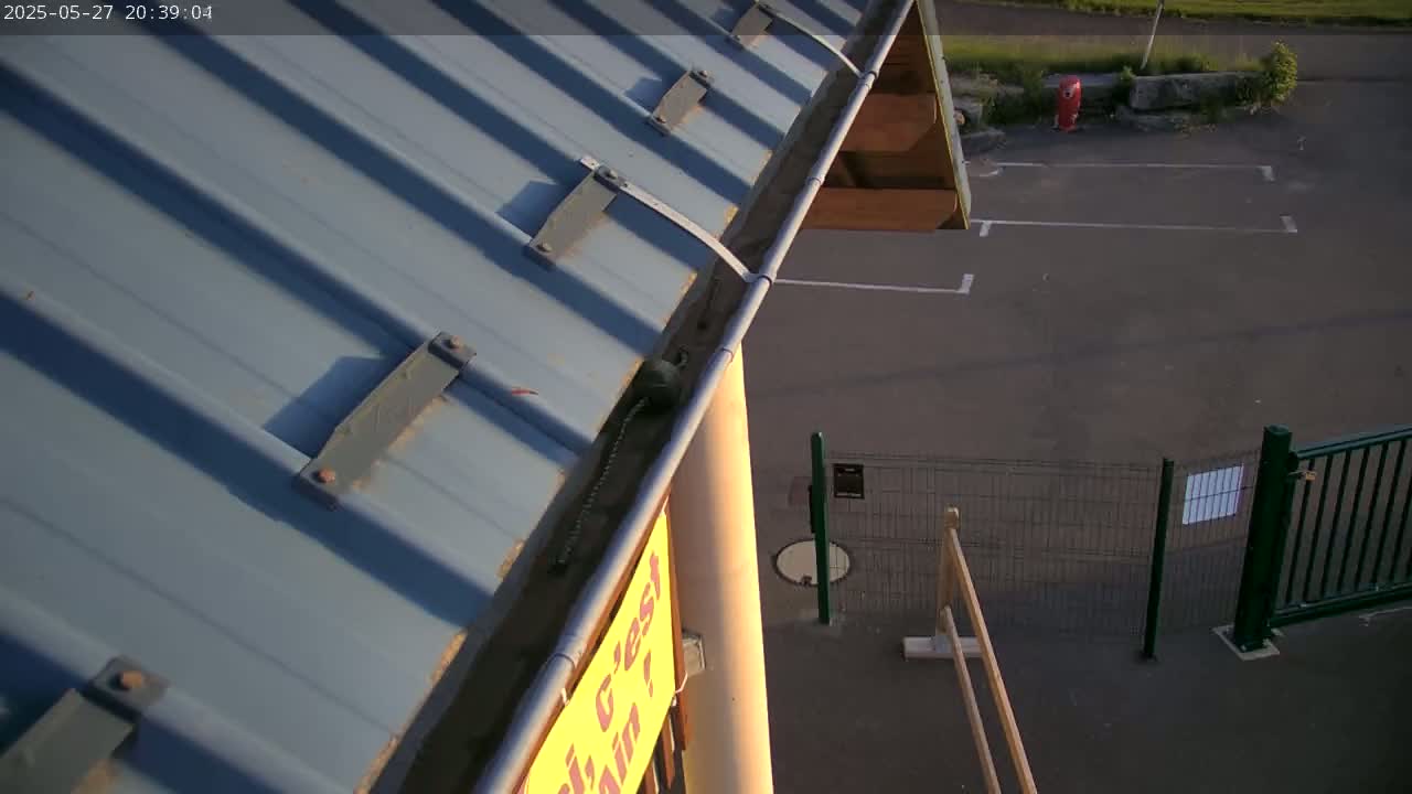 A high-angle view shows a blue metal roof with a gutter, alongside a paved area enclosed by a metal fence, under partly sunny evening conditions.