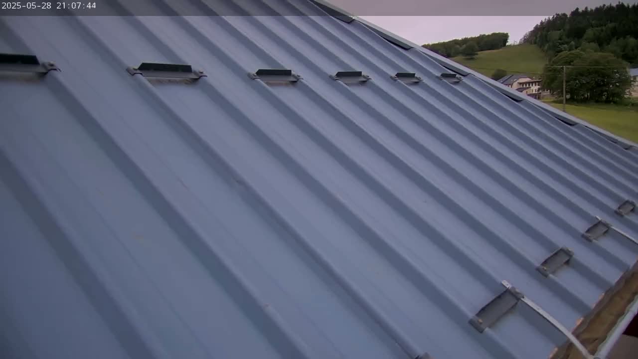 A light-blue corrugated metal roof with several rectangular vents is angled towards the viewer, overlooking a partly cloudy sky and distant green hills with a few buildings.