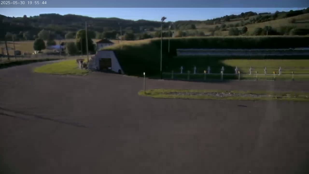 A paved area curves around a grassy field with a partially subterranean building and a line of evenly spaced markers under a clear sky.