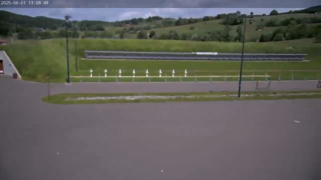 An overcast day shows a paved area in front of a shooting range with targets, set against a backdrop of rolling green hills.